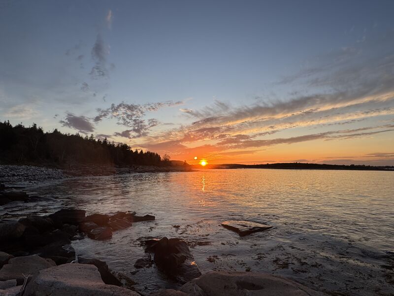 The image captures a serene sunset over a body of water, likely a lake or ocean. The sky is painted with hues of orange and blue, reflecting on the calm water. Silhouetted trees line the left side, and rocks are in the foreground. The overall scene evokes a sense of peace and tranquility.